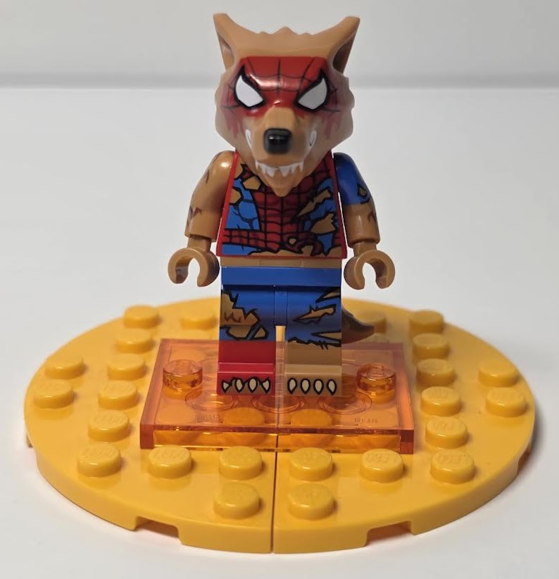 May include: A Lego minifigure of a wolf-like creature with a red and brown mask, brown arms, and a torn blue and red outfit. The figure stands on a yellow Lego base with an orange transparent platform.