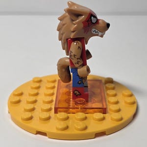 May include: A Lego minifigure of a werewolf with a brown fur head, red shirt, and blue pants. The werewolf has a tail and is standing on a yellow Lego base. The minifigure has a red spiderweb design on its face.