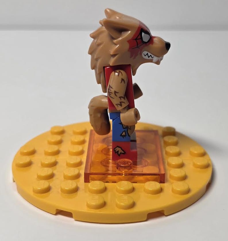 May include: A Lego minifigure of a werewolf with a brown fur head, red shirt, and blue pants. The werewolf has a tail and is standing on a yellow Lego base. The minifigure has a red spiderweb design on its face.