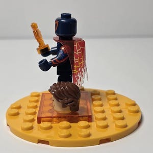 Puede incluir: Una minifigura de Lego con una cabeza azul oscuro y una capa estampada en rojo y blanco, sosteniendo un accesorio dorado. La figura est&aacute; sobre una base circular amarilla con una segunda cabeza y un soporte naranja transparente.