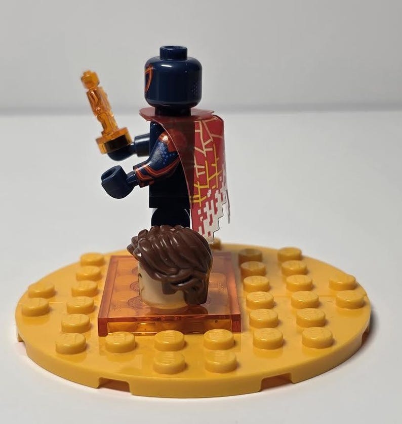 Puede incluir: Una minifigura de Lego con una cabeza azul oscuro y una capa estampada en rojo y blanco, sosteniendo un accesorio dorado. La figura est&aacute; sobre una base circular amarilla con una segunda cabeza y un soporte naranja transparente.