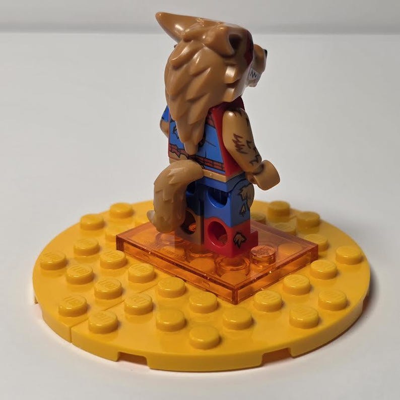 May include: A Lego minifigure of a werewolf with brown fur, a red vest, and blue pants. The figure stands on a clear orange base, which is set on a yellow Lego baseplate. The figure has a tail and arm tattoos.