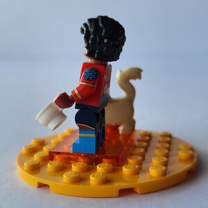Puede incluir: Una minifigura de Lego con una peluca rizada negra, una chaqueta roja y pantalones azules est&aacute; de pie sobre una base amarilla. La figura sostiene un objeto blanco y est&aacute; junto a una figura de animal de color beige. La base es un c&iacute;rculo amarillo con clavijas amarillas.