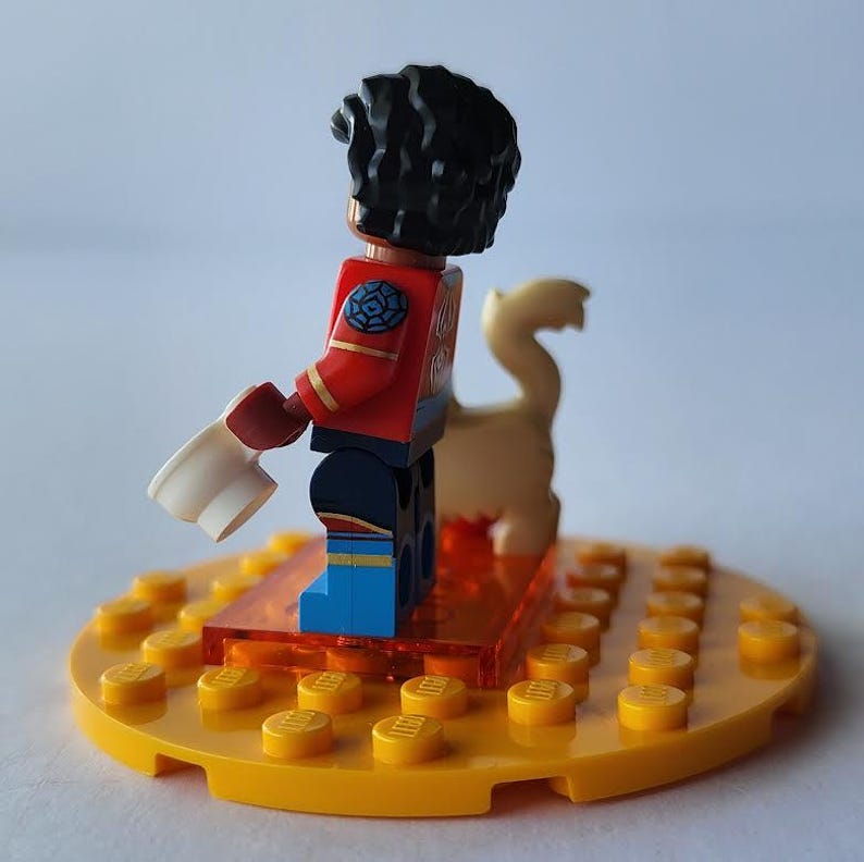 Puede incluir: Una minifigura de Lego con una peluca rizada negra, una chaqueta roja y pantalones azules est&aacute; de pie sobre una base amarilla. La figura sostiene un objeto blanco y est&aacute; junto a una figura de animal de color beige. La base es un c&iacute;rculo amarillo con clavijas amarillas.