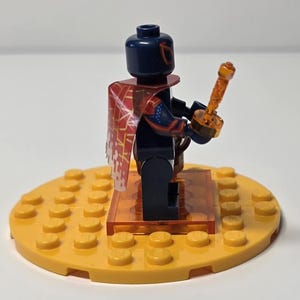 Puede incluir: Una minifigura de Lego con cabeza y torso azul oscuro, que lleva una capa roja y blanca. La figura sostiene una espada naranja y se encuentra sobre una base circular amarilla. La base est&aacute; hecha de ladrillos Lego.