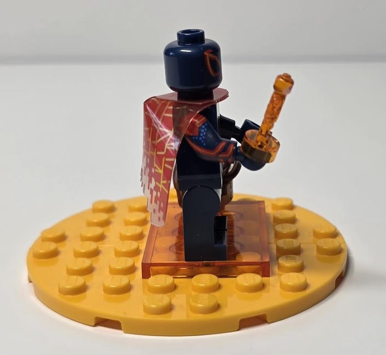 Puede incluir: Una minifigura de Lego con cabeza y torso azul oscuro, que lleva una capa roja y blanca. La figura sostiene una espada naranja y se encuentra sobre una base circular amarilla. La base est&aacute; hecha de ladrillos Lego.
