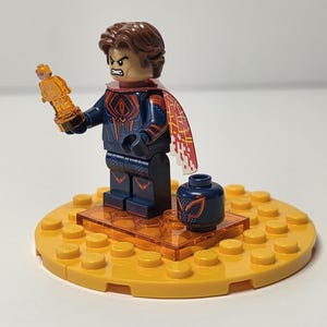 Puede incluir: Una minifigura de Lego con cabello casta&ntilde;o, un traje azul y rojo y una capa roja y blanca. La figura sostiene un trofeo dorado y est&aacute; de pie sobre una base amarilla con un peque&ntilde;o recipiente azul.