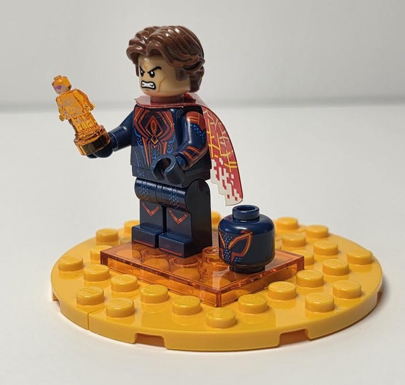 Puede incluir: Una minifigura de Lego con cabello casta&ntilde;o, un traje azul y rojo y una capa roja y blanca. La figura sostiene un trofeo dorado y est&aacute; de pie sobre una base amarilla con un peque&ntilde;o recipiente azul.