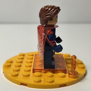 Puede incluir: Una minifigura de Lego con cabello casta&ntilde;o y una capa estampada en rojo y dorado. La figura est&aacute; de pie sobre una base Lego amarilla con un peque&ntilde;o trofeo dorado. La figura lleva un traje azul y rojo con guantes negros.