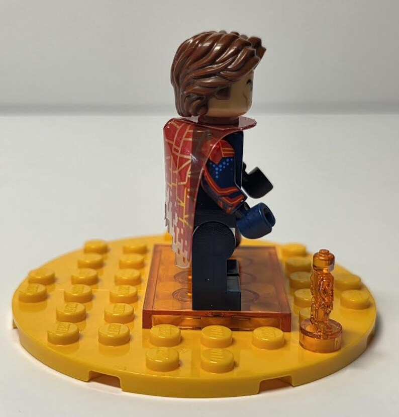 Puede incluir: Una minifigura de Lego con cabello casta&ntilde;o y una capa estampada en rojo y dorado. La figura est&aacute; de pie sobre una base Lego amarilla con un peque&ntilde;o trofeo dorado. La figura lleva un traje azul y rojo con guantes negros.