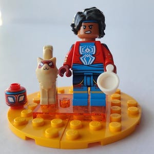 Puede incluir: Una minifigura de Lego de una persona con un atuendo rojo y azul, sosteniendo un objeto blanco, de pie sobre una base amarilla. Una figura de gato blanco y una cabeza roja y azul tambi&eacute;n est&aacute;n en la base. La minifigura tiene cabello negro y una diadema azul.