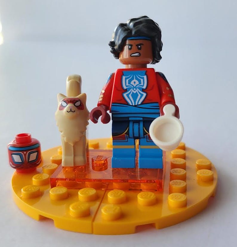 Puede incluir: Una minifigura de Lego de una persona con un atuendo rojo y azul, sosteniendo un objeto blanco, de pie sobre una base amarilla. Una figura de gato blanco y una cabeza roja y azul tambi&eacute;n est&aacute;n en la base. La minifigura tiene cabello negro y una diadema azul.