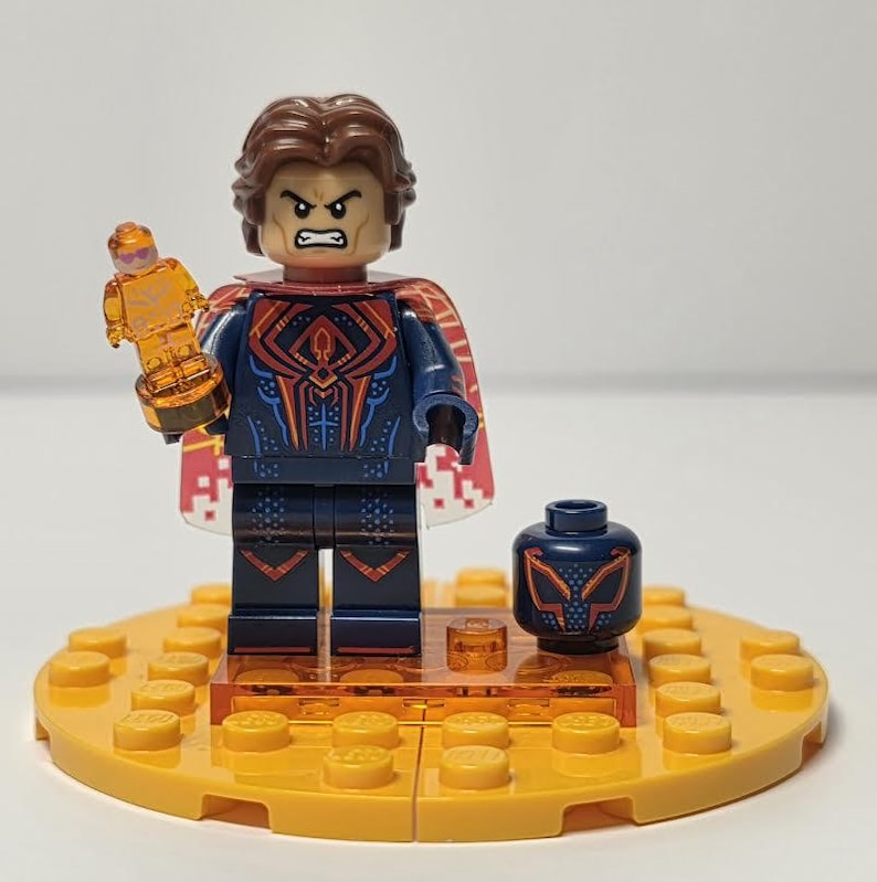 Puede incluir: Una minifigura de Lego con cabello casta&ntilde;o y una expresi&oacute;n severa, con un disfraz azul y rojo, sosteniendo un trofeo dorado. La figura est&aacute; sobre una base amarilla con piezas de Lego adicionales y una cabeza de repuesto.