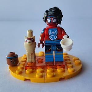 Puede incluir: Una minifigura de Lego de un personaje con un traje rojo y azul con un emblema blanco, sosteniendo una taza blanca. Un gato Lego y una peque&ntilde;a figura Lego marr&oacute;n con una cara sonriente tambi&eacute;n est&aacute;n presentes en una base Lego amarilla.