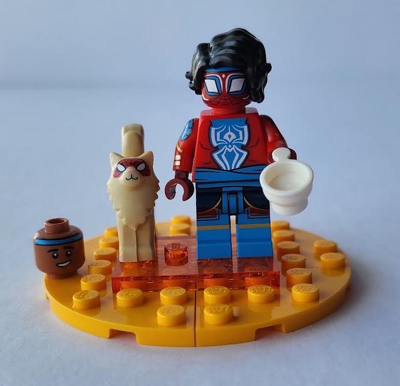 Puede incluir: Una minifigura de Lego de un personaje con un traje rojo y azul con un emblema blanco, sosteniendo una taza blanca. Un gato Lego y una peque&ntilde;a figura Lego marr&oacute;n con una cara sonriente tambi&eacute;n est&aacute;n presentes en una base Lego amarilla.