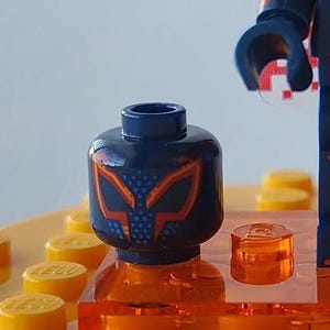 Puede incluir: Primer plano de una cabeza de minifigura de Lego con un casco azul oscuro con un dise&ntilde;o de m&aacute;scara naranja y azul. La cabeza se asienta sobre un ladrillo Lego naranja, rodeado de clavijas Lego amarillas. Se ve una parte del brazo de la minifigura.