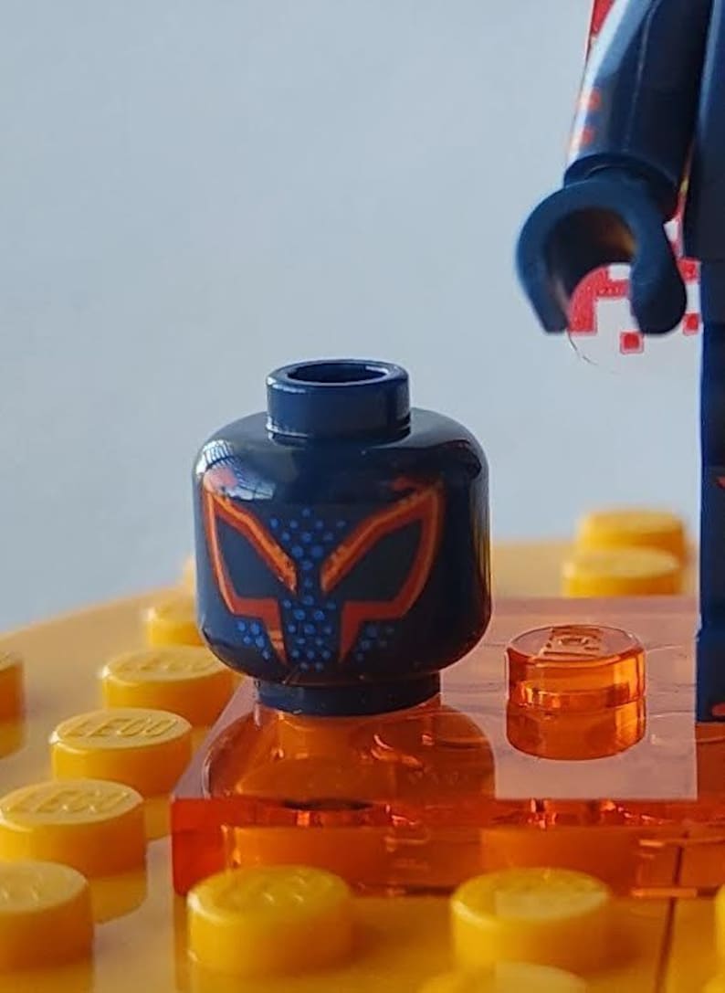 Puede incluir: Primer plano de una cabeza de minifigura de Lego con un casco azul oscuro con un dise&ntilde;o de m&aacute;scara naranja y azul. La cabeza se asienta sobre un ladrillo Lego naranja, rodeado de clavijas Lego amarillas. Se ve una parte del brazo de la minifigura.