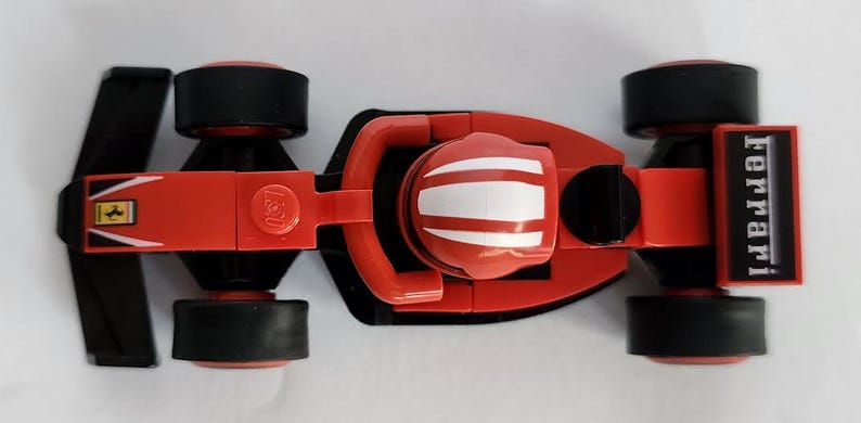 May include: A red toy race car with black wheels and a white stripe on the helmet. The car has a "Ferrari" logo on the side and a "Lego" logo on the bottom.
