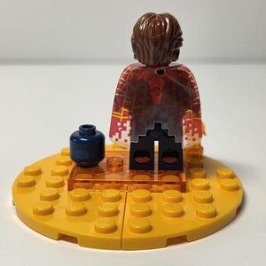 Puede incluir: Una minifigura de Lego con cabello casta&ntilde;o y una capa estampada en rojo y blanco se encuentra sobre una base Lego amarilla. Un accesorio Lego azul oscuro con puntos blancos est&aacute; en la base. Una peque&ntilde;a pieza Lego transparente est&aacute; frente a la figura.