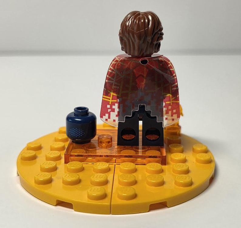 Puede incluir: Una minifigura de Lego con cabello casta&ntilde;o y una capa estampada en rojo y blanco se encuentra sobre una base Lego amarilla. Un accesorio Lego azul oscuro con puntos blancos est&aacute; en la base. Una peque&ntilde;a pieza Lego transparente est&aacute; frente a la figura.