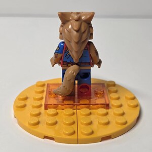 May include: A Lego minifigure with a wolf-like head and tail, wearing a blue and red outfit. The figure stands on a clear orange platform, which is set on a yellow Lego base. The figure has brown hair and a tan tail.