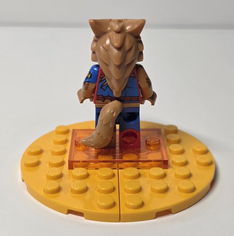 May include: A Lego minifigure with a wolf-like head and tail, wearing a blue and red outfit. The figure stands on a clear orange platform, which is set on a yellow Lego base. The figure has brown hair and a tan tail.