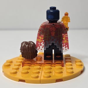 Puede incluir: Una minifigura de Lego con una cabeza azul oscuro y una capa estampada en rojo y blanco. La figura sostiene una peque&ntilde;a figura naranja transl&uacute;cida. Un mech&oacute;n de pelo casta&ntilde;o y una base Lego amarilla completan la escena.