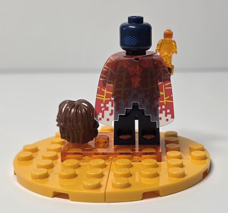 Puede incluir: Una minifigura de Lego con una cabeza azul oscuro y una capa estampada en rojo y blanco. La figura sostiene una peque&ntilde;a figura naranja transl&uacute;cida. Un mech&oacute;n de pelo casta&ntilde;o y una base Lego amarilla completan la escena.