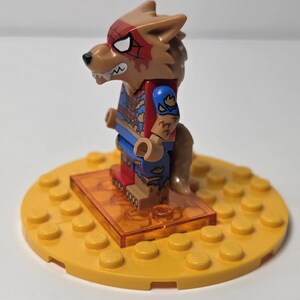May include: A Lego minifigure of a werewolf character, featuring a brown wolf head with red spiderweb markings and white eyes. The figure wears a blue and red costume and stands on a yellow base with a clear orange stand.