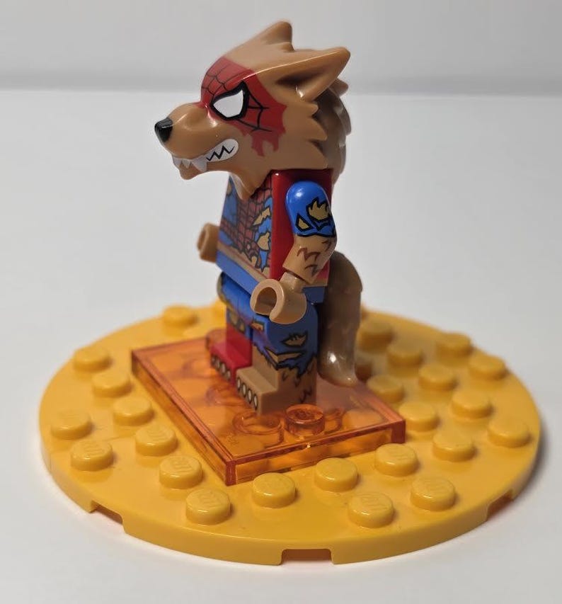 May include: A Lego minifigure of a werewolf character, featuring a brown wolf head with red spiderweb markings and white eyes. The figure wears a blue and red costume and stands on a yellow base with a clear orange stand.