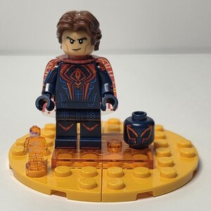 Puede incluir: Una minifigura de Lego de un superh&eacute;roe con un postizo marr&oacute;n, un traje azul oscuro con detalles rojos y naranjas, y una capa roja. La figura est&aacute; sobre una base amarilla con piezas de Lego adicionales y una cabeza de repuesto.
