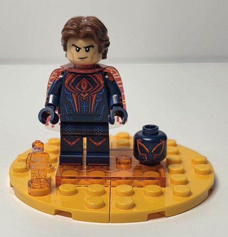 Puede incluir: Una minifigura de Lego de un superh&eacute;roe con un postizo marr&oacute;n, un traje azul oscuro con detalles rojos y naranjas, y una capa roja. La figura est&aacute; sobre una base amarilla con piezas de Lego adicionales y una cabeza de repuesto.