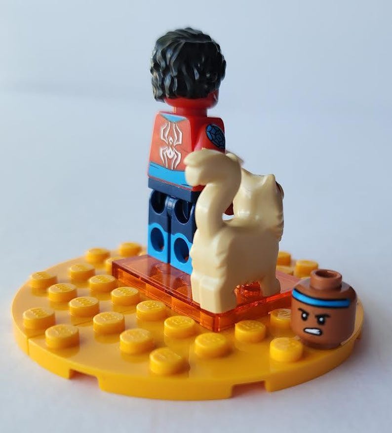 Puede incluir: Una minifigura de Lego con torso rojo y azul y pelo negro est&aacute; sobre una base amarilla. Una figura de animal de color canela y una olla marr&oacute;n con una franja azul tambi&eacute;n est&aacute;n en la base. La minifigura tiene un dise&ntilde;o de ara&ntilde;a blanca en el pecho.