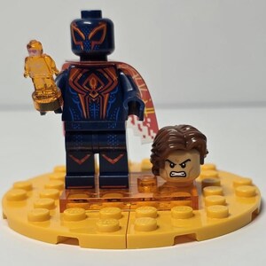Puede incluir: Una minifigura de Lego de un superh&eacute;roe con un traje azul oscuro con detalles rojos y naranjas, sosteniendo un trofeo dorado. La figura tiene una m&aacute;scara y una capa a juego. Tambi&eacute;n se incluye una segunda cabeza con cabello casta&ntilde;o y expresi&oacute;n enfadada.