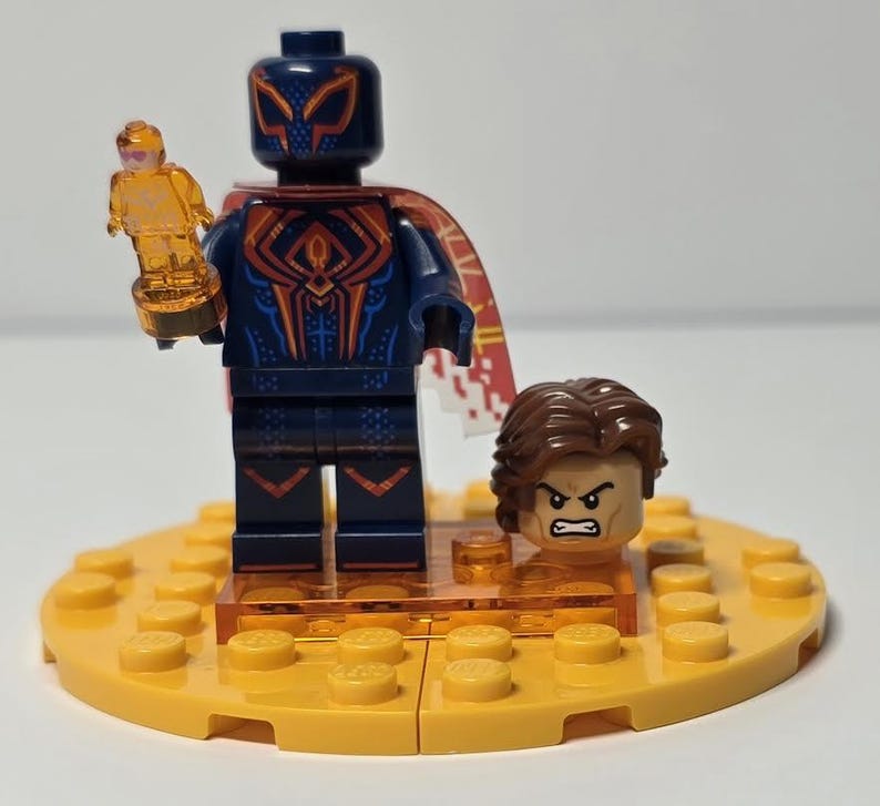 Puede incluir: Una minifigura de Lego de un superh&eacute;roe con un traje azul oscuro con detalles rojos y naranjas, sosteniendo un trofeo dorado. La figura tiene una m&aacute;scara y una capa a juego. Tambi&eacute;n se incluye una segunda cabeza con cabello casta&ntilde;o y expresi&oacute;n enfadada.
