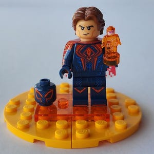 Puede incluir: Una minifigura de Lego de un personaje con un traje azul y rojo, sosteniendo una peque&ntilde;a figura naranja. La minifigura est&aacute; sobre una base amarilla con detalles en naranja y una cabeza azul separada con un dise&ntilde;o de m&aacute;scara.