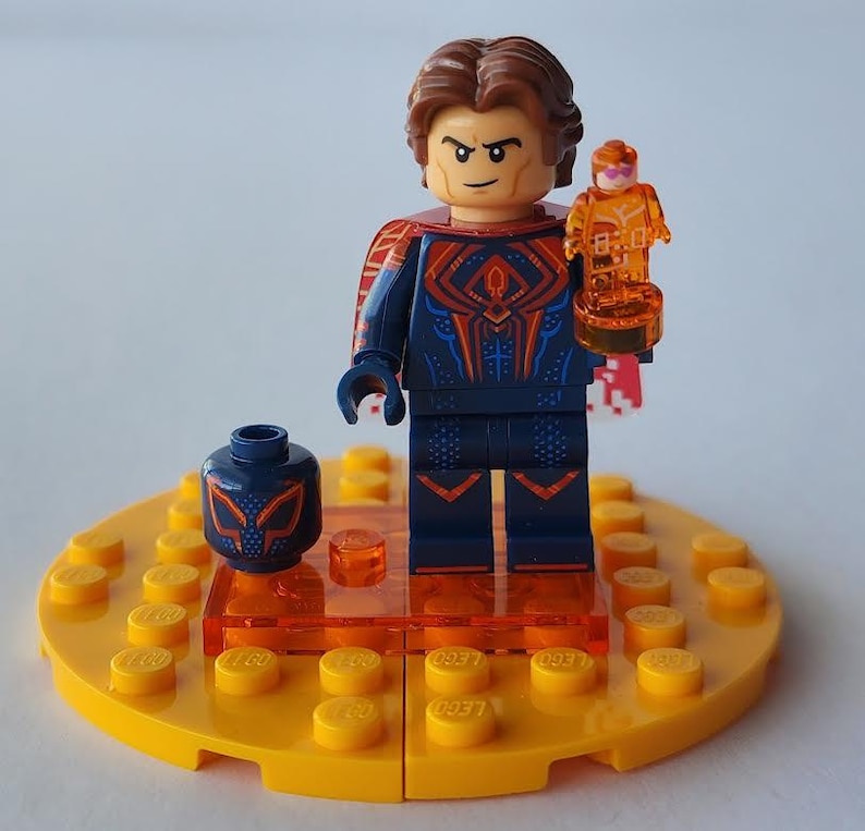 Puede incluir: Una minifigura de Lego de un personaje con un traje azul y rojo, sosteniendo una peque&ntilde;a figura naranja. La minifigura est&aacute; sobre una base amarilla con detalles en naranja y una cabeza azul separada con un dise&ntilde;o de m&aacute;scara.