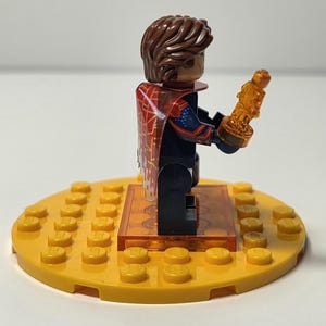 Puede incluir: Una minifigura de Lego con cabello casta&ntilde;o y una capa roja y dorada, sosteniendo un trofeo dorado. La figura est&aacute; sobre una base Lego amarilla con una plataforma naranja transparente. Es un juguete.