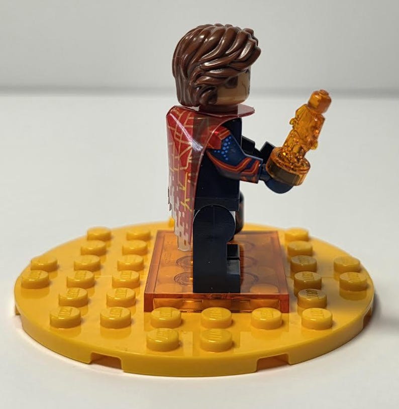 Puede incluir: Una minifigura de Lego con cabello casta&ntilde;o y una capa roja y dorada, sosteniendo un trofeo dorado. La figura est&aacute; sobre una base Lego amarilla con una plataforma naranja transparente. Es un juguete.