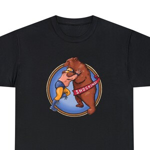 May include: Black t-shirt with a vintage illustration of a wrestler grappling with a brown bear. The illustration is surrounded by a blue circle with a gold frame and a red banner that reads "SHOCKING!"