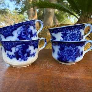 May include: Four antique teacups with matching saucers. The cups are white with a blue floral pattern and a blue rim. The cups are sitting on a wooden surface.
