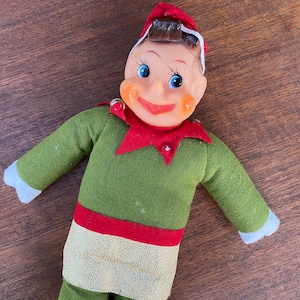 May include: A vintage elf doll with a plastic face and a green felt outfit. The elf has a red collar and hat, with white trim on the hat and sleeves. The doll's face has blue eyes and a smiling expression.