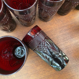 May include: A collection of ruby red, cut-glass tumblers with intricate geometric and floral patterns. The glasses are arranged on a wooden surface, showcasing their detailed designs and reflective qualities.