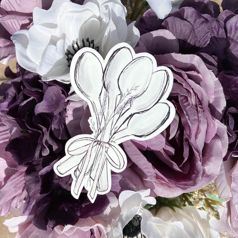 May include: A white sticker with a black outline of a bouquet of spoons. The sticker is placed on a background of purple and white flowers.