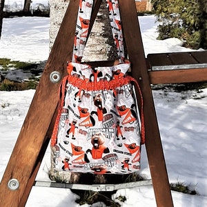 May include: A white and orange fabric tote bag with a cartoon character design hanging on a wooden ladder. The bag has a drawstring closure and a short handle.