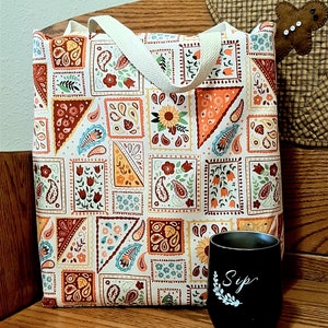 May include: A white tote bag with a colorful paisley pattern. The bag has two white canvas straps. The bag is sitting on a wooden surface next to a black mug with the word "Sip" written on it in white.