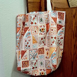 May include: A white tote bag with a patchwork design featuring paisley patterns, flowers, and geometric shapes in shades of orange, red, yellow, and blue.