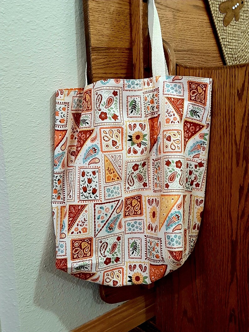 May include: A white tote bag with a patchwork design featuring paisley patterns, flowers, and geometric shapes in shades of orange, red, yellow, and blue.