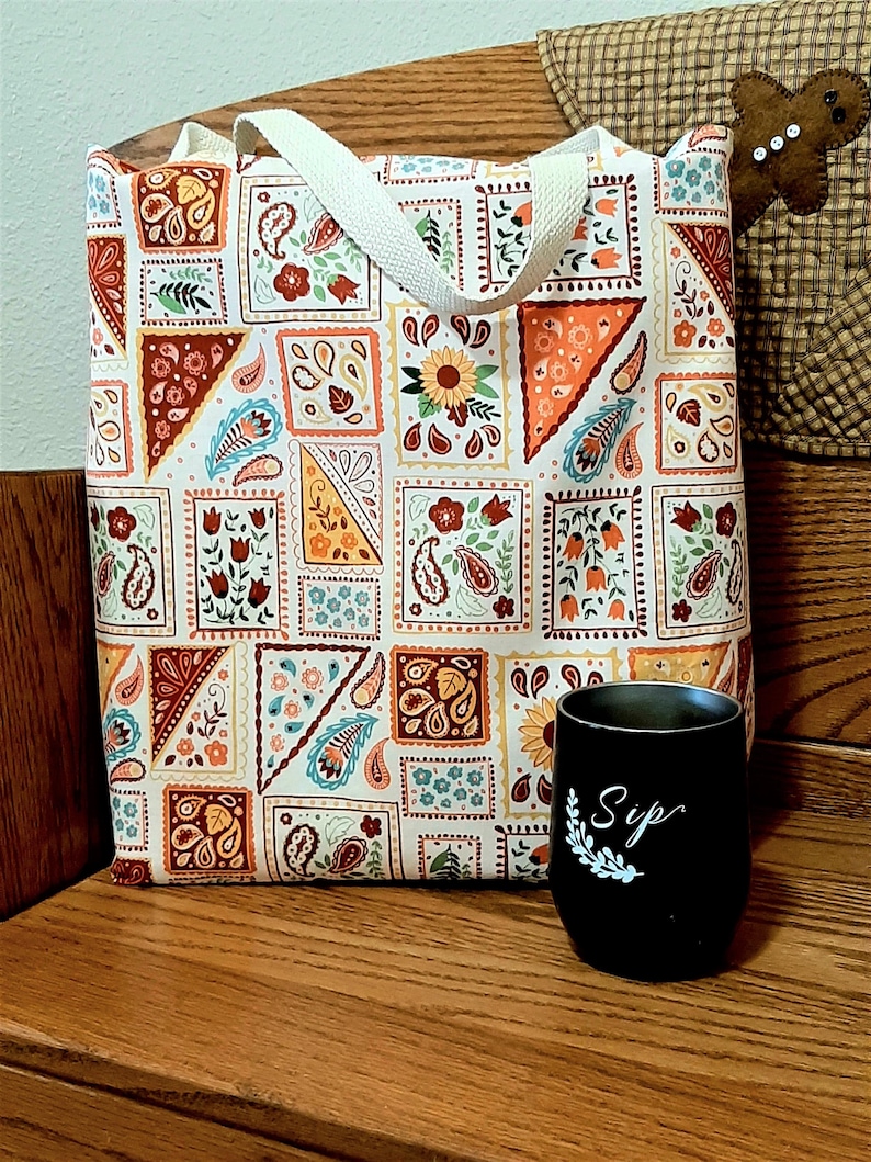 May include: A white tote bag with a colorful paisley pattern. The bag has two white canvas straps. The bag is sitting on a wooden surface next to a black mug with the word "Sip" written on it in white.