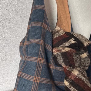 May include: A hobo-style shoulder bag made of blue fabric with an orange plaid pattern, featuring a wide strap and a tan interior lining. A folded scarf with a brown, cream, and dark red plaid pattern is tucked inside the bag's main opening.